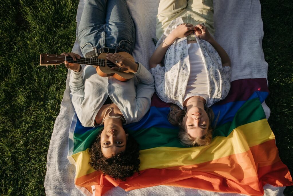 Diverse friends enjoy a sunny day lying on a rainbow blanket, one playing ukulele.